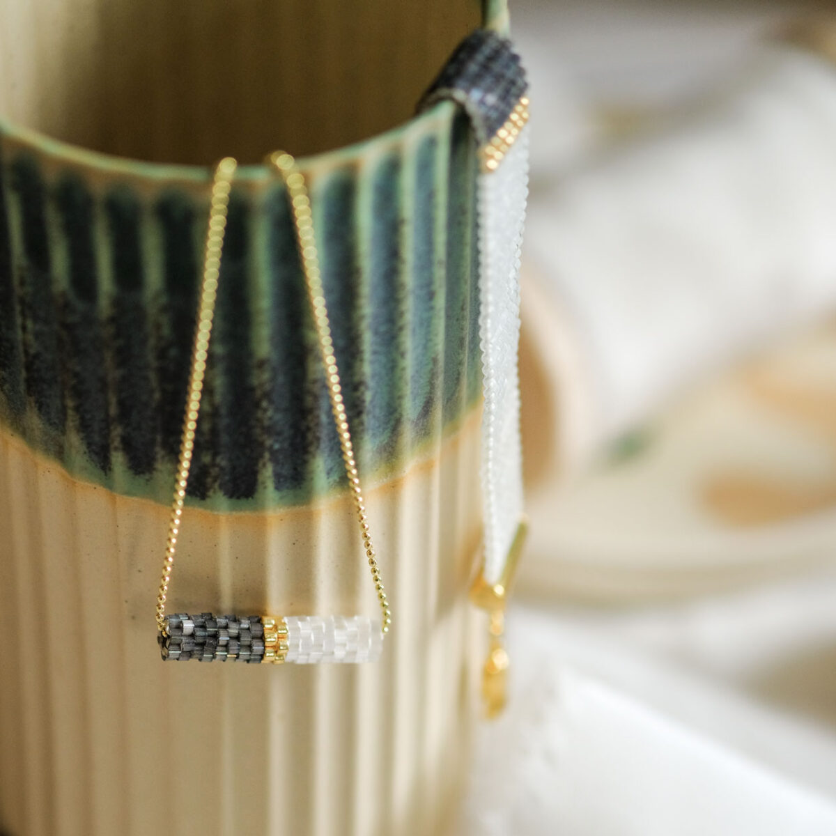 Elegant handcrafted bead necklace and gold chain displayed on a ceramic vase with green brush strokes.
