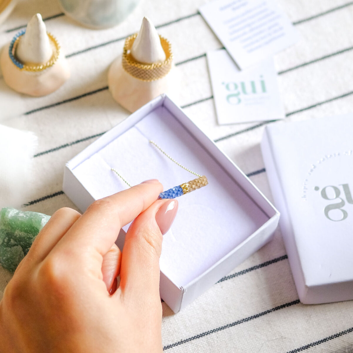A hand holding a delicate blue, beige and gold bar necklace above an open jewelry box, with branding materials in the background.
